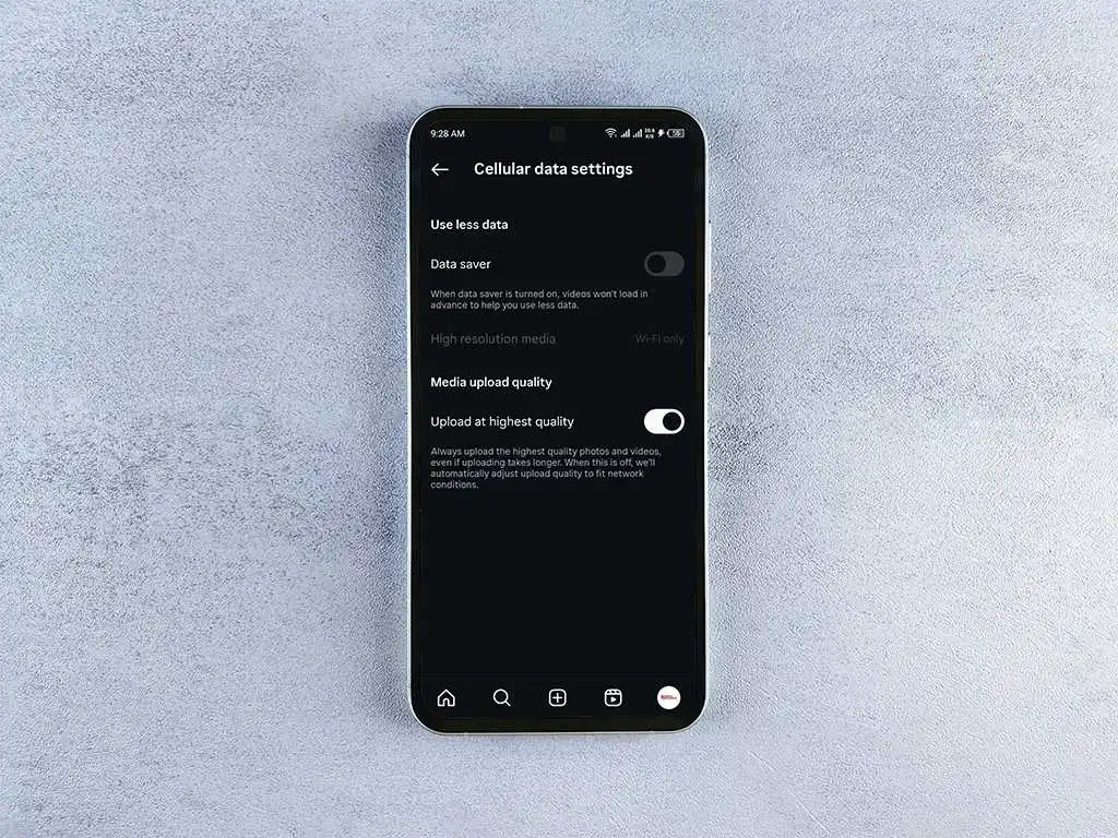 A screenshot of a phone in dark mode showing the "Cellular data settings" page within the Instagram app. Options displayed include "Data saver" and "Upload at highest quality," both with toggle switches, along with a "High resolution media" setting for "Wi-fi only." The phone is held against a light grey surface.